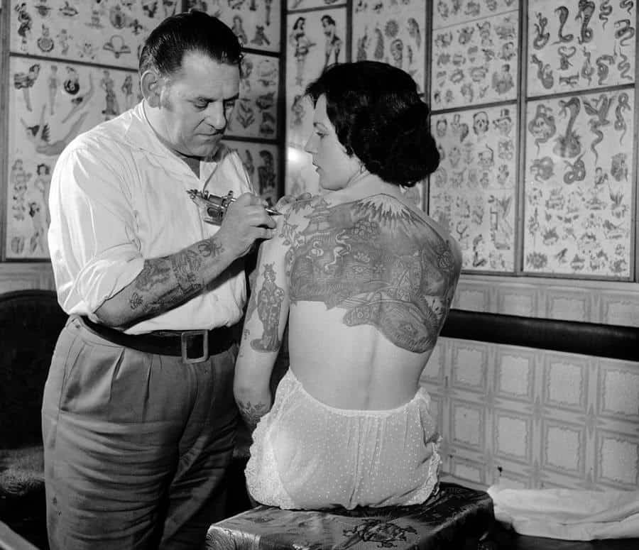 25. (سكوز) وهو يقوم بوشم لبطلة الوشوم (بام ناش) في عام 1960.