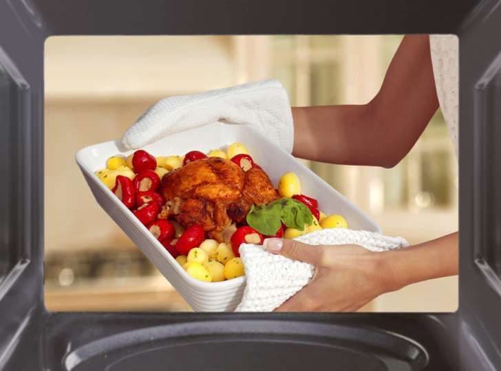 صورة امرأة تحمل إناء طهو مع لحم وبطاطس أمام جهاز مايكرويف