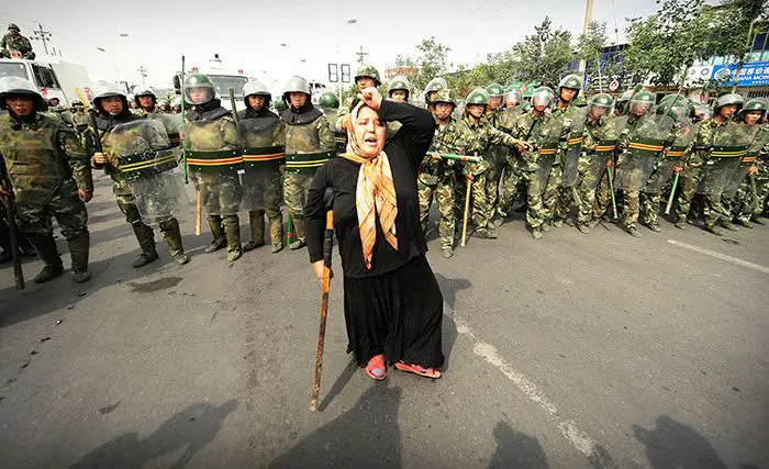 شرطة مكافحة الشغب الصينية تراقب احتجاجاً لامرأة من الاثنية اليوغورية في (أورومتشي) Urumiqi في أقصى غرب الصين في مقاطعة (شينجيانغ) Xinjiang، بعد ثالث يوم من الإضرابات، بتاريخ 7 تموز/يوليو 2009.