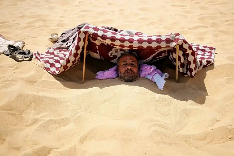 أحد "المرضى" الذين تم دفنه في الرمال الساخنة يراقب من تحت ملاءة تلقي عليه بالظلال لحماية وجهه ورأسه من حرارة الشمس اللاذعة في (سيوا)، بمصر.