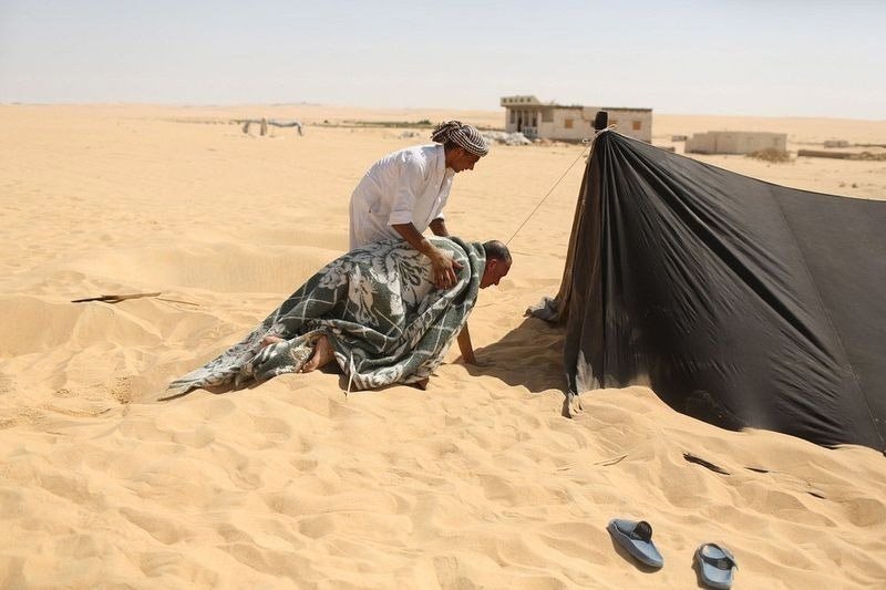عامل يساعد أحد المرضى الملتحف ملاءة للذهاب إلى خيمة السونا بعد أن أٌخرج من الرمال الساخنة التي كان مدفونا فيها في (سيوا) بمصر.