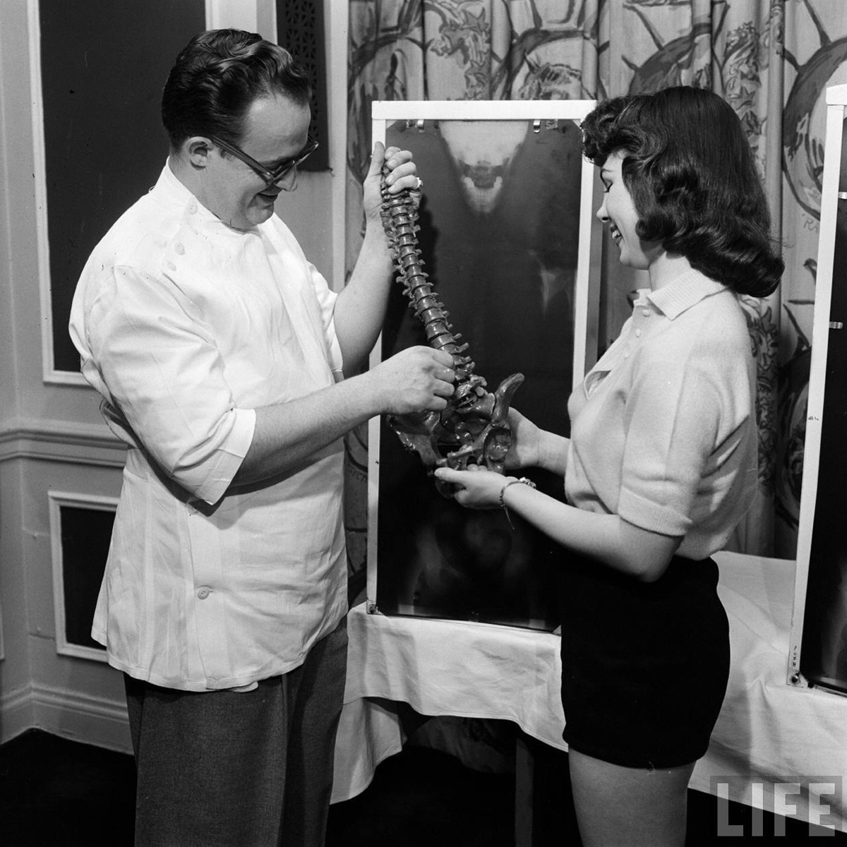 المتسابقة (لويس كونواي) وهي تقف بجانب صورة سينية لعمودها الفقري بينما يقوم ممارس لعلاج تقويم العظام بحمل مجسم للعمود الفقري البشري ليقارنه بالصورة، التقطت في مايو 1946 - صورة: Wallace Kirkland/LIFE