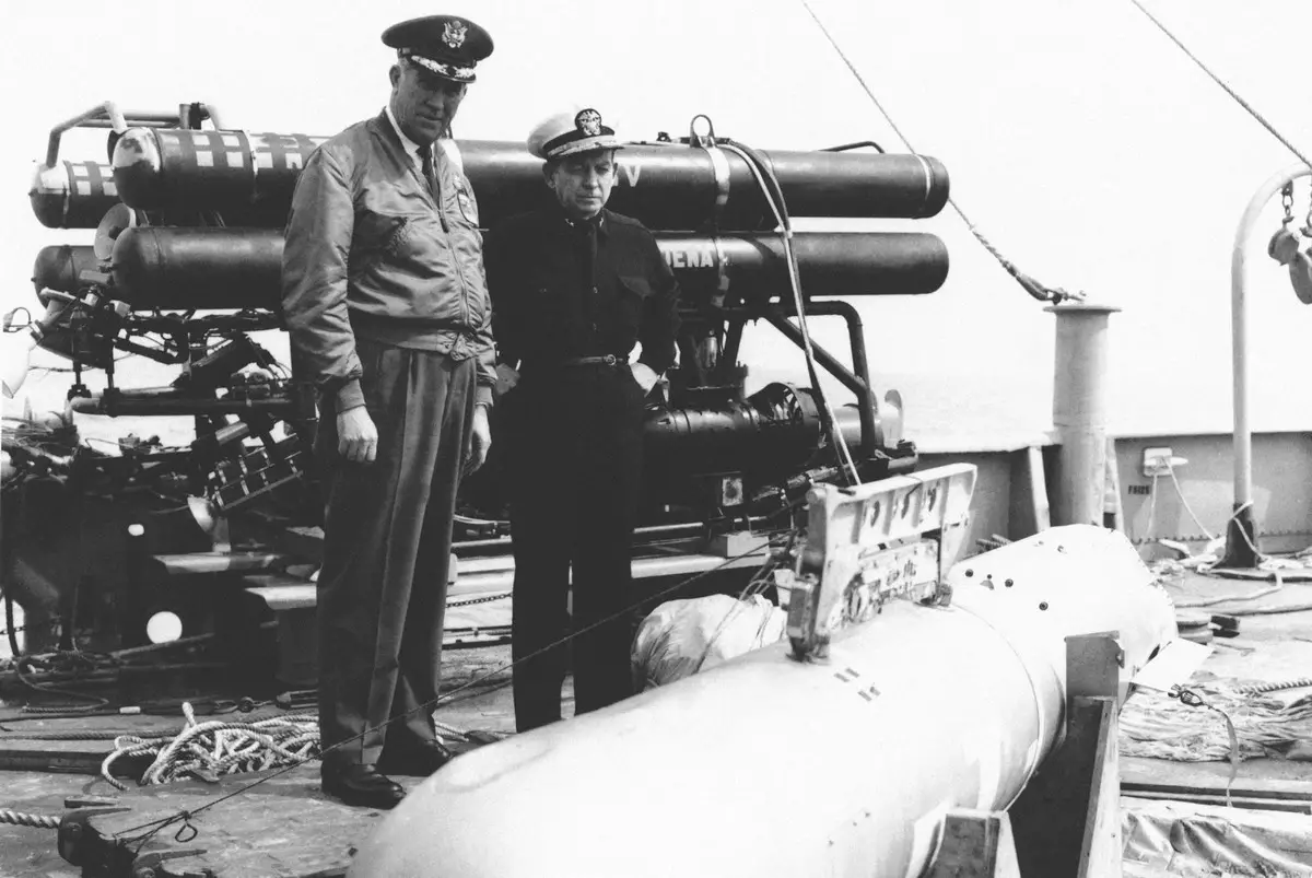 جنرال القوات الجوية الأمريكية (ديلمار ويلسن) على اليسار، وأدميرال القوات البحرية الأمريكية (ويليام غيست) على اليمين يتفحصان القنبلة النووية الرابعة التي عثر عليها في عرض البحر بعد استخراجها.