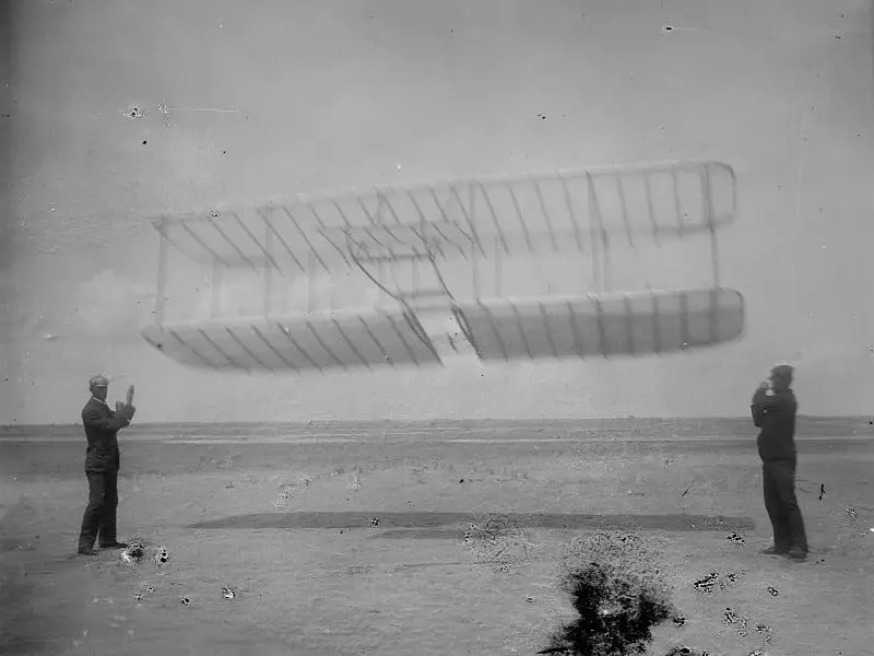 من اليسار إلى اليمين: (ويلبير)، طائرة (ذا رايت)، و(أورفيل) شقيقه، في (كيتي هاوك) في كارولاينا الشمالية سنة 1901.